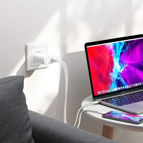 Hvit, kompakt USB-C-vegglader i stikkontakt, to porter med to hvite kabler som lader en bærbar datamaskin og en smarttelefon på bordet.