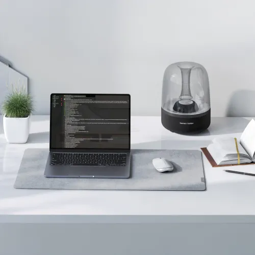 ALOGIC Echelon balta žemo profilio belaidė pelė ant pilko stalinio kilimėlio, padėta į dešinę nuo nešiojamojo kompiuterio ir rašiklio.