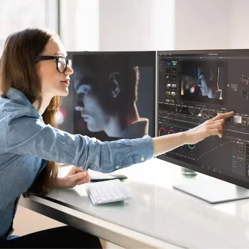 32-tums pekskärmsmonitor på silverstativ, tunn ram, på skrivbord; en hand pekar på skärmen med videoredigeringsgränssnitt.