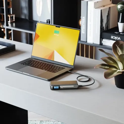 ALOGIC Ruck powerbank i metall med liten statusdisplay, ansluten med USB-C-kabel till laptop på skrivbord bredvid krukväxt.