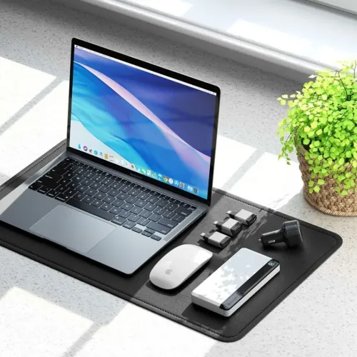 USB-C uz DisplayPort adapteris pelēkā metāla krāsā ar USB-C krānu un taisnstūra portu, uz melna statīva blakus klēpjdatoram.