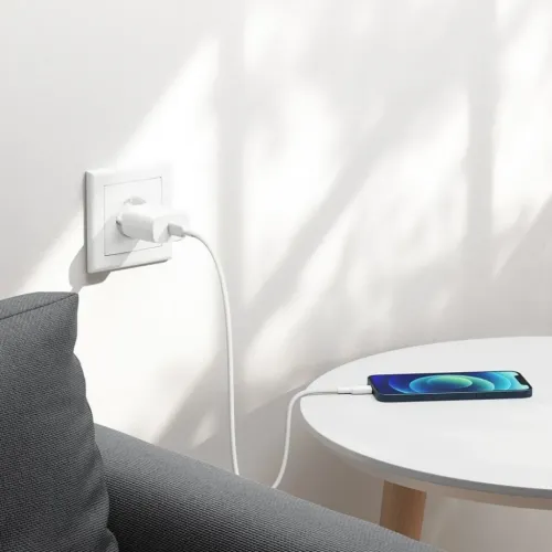 USB-C-väggladdare i vitt, ansluten till vägguttag med vit kabel som når en telefon på rund vit sidobord intill grå soffa.