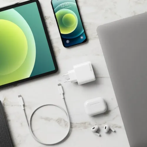 Vit fyrkantig väggladdare med EU-stickpropp och USB‑C‑kabel på marmoryta intill surfplatta, telefon, laptop och hörlurar