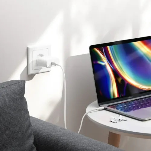 USB‑C-laddare i vitt, ansluten i vägguttag, med lång kabel till bärbar dator på runt sidobord; soffa och hörlurar intill.