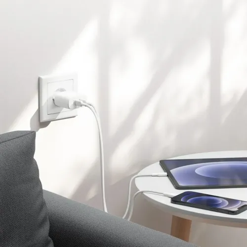 Kompakts balts USB-C sienas lādētājs ar diviem kabeļiem, kas savienots ar sienas kontaktligzdu, uzlādē planšetdatoru un tālruni uz apaļas baltas galda virsmas.