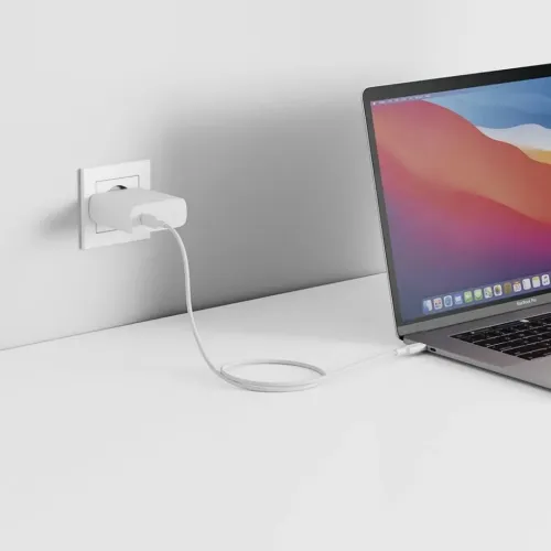 Valkoinen, suorakulmainen USB-C-seinälaturi istuu seinäpistorasiaan; kaapeli kulkee pöydän yli kannettavaan tietokoneeseen oikealla.