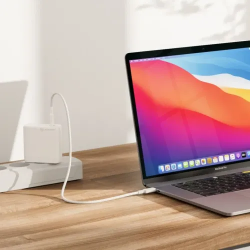 Valkoinen ALOGIC 100W GaN -laturi USB-C-kaapelilla, joka on kytketty pistorasiaan ja kannettavaan tietokoneeseen puupöydällä, sivulta katsottuna.