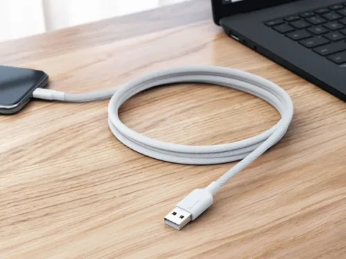 Hvit, flettet USB-A til USB-C-kabel, delvis opprullet på et trebord, koblet til en mobiltelefon med USB-C; USB-A-enden mot kameraet.
