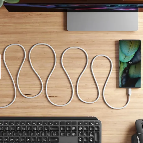 White silicone USB-C to USB-C cable lies wave-shaped on a wooden table and connects to a smartphone charging on the table.