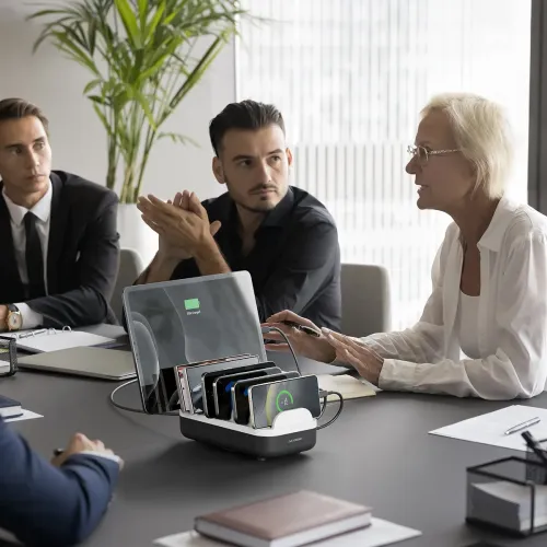 USB-C skrivbordsladdningsstation med flera fack, telefoner och en surfplatta dockade med kablar på konferensbord i möte.