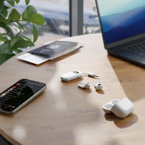 Hvid Bluetooth-lydsender med display, knapper og 3,5 mm adapterstik på træbord ved siden af hovedtelefoner, mobiltelefon og laptop.