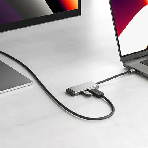 Sidabriškai pilkas "3-in-1" USB-C adapteris su HDMI ir USB-A prievadais, jungiamas prie nešiojamojo kompiuterio ir išorinio monitoriaus ant ryškaus darbastalio.